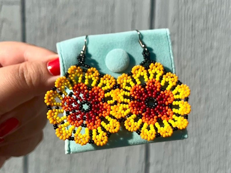 K&ouml;nnte beinhalten: Ein Paar Perlenohrringe mit Blumenmuster. Die Ohrringe sind aus schwarzen, roten, orangefarbenen und gelben Perlen gefertigt. Die Ohrringe befinden sich auf einem hellblauen Hintergrund.