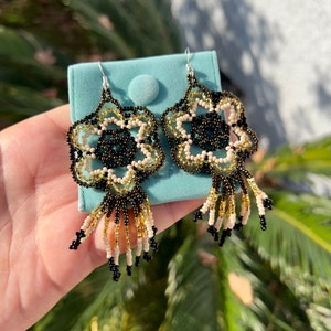 May include: A pair of beaded earrings with a floral design in black, gold, and cream colors. The earrings have a fringe of beads hanging from the bottom.