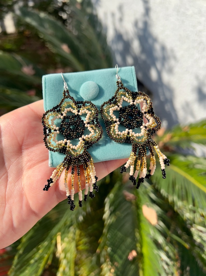 May include: A pair of beaded earrings with a floral design in black, gold, and cream colors. The earrings have a fringe of beads hanging from the bottom.