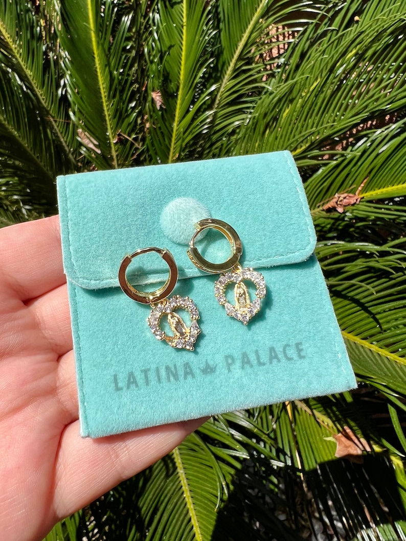 Puede incluir: Pendientes de aro dorados con colgantes en forma de coraz&oacute;n. Cada colgante presenta una figura religiosa central rodeada de cristales transparentes. Los pendientes se muestran en una bolsa de terciopelo azul claro con el texto "LATINA PALACE".