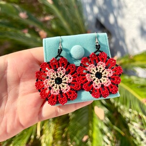 K&ouml;nnte beinhalten: Ein Paar rote, rosa und schwarze Perlen-Blumenohrringe. Die Ohrringe h&auml;ngen an silbernen Haken und werden auf einem blauen Samtbeutel pr&auml;sentiert.