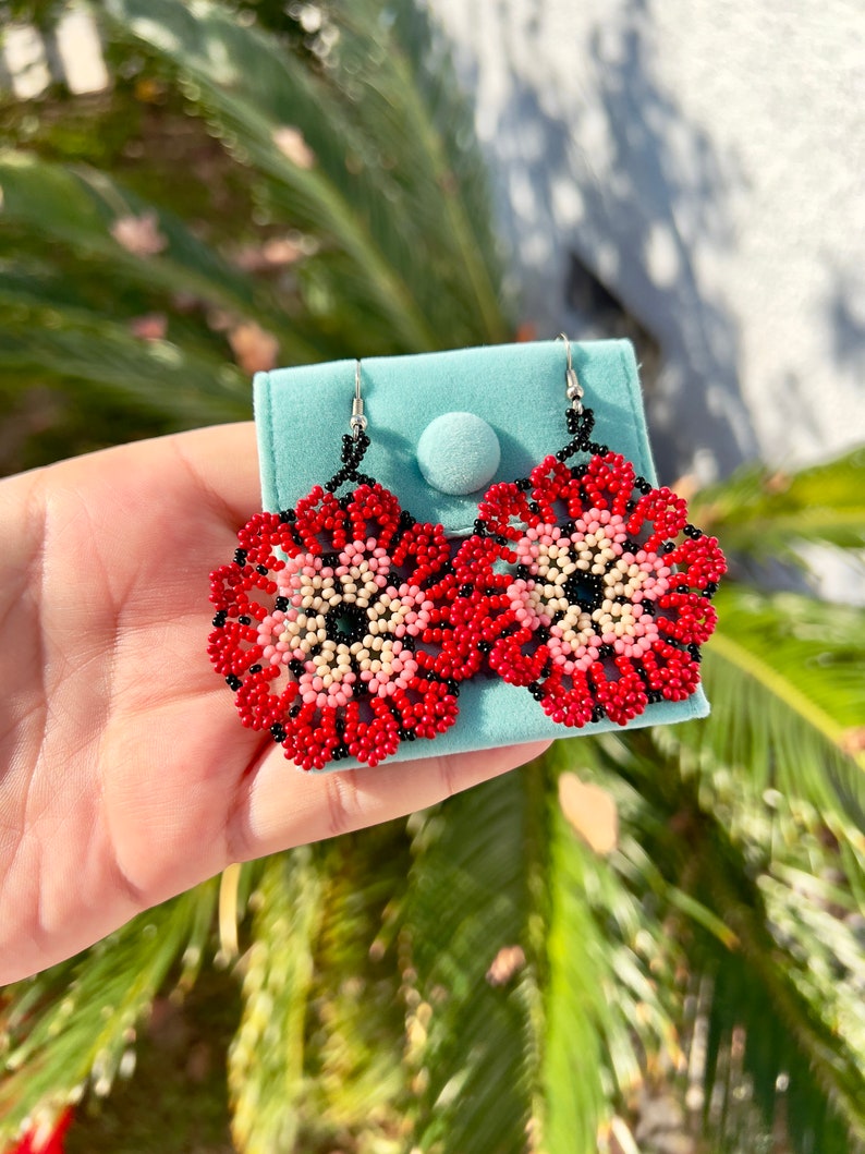 K&ouml;nnte beinhalten: Ein Paar rote, rosa und schwarze Perlen-Blumenohrringe. Die Ohrringe h&auml;ngen an silbernen Haken und werden auf einem blauen Samtbeutel pr&auml;sentiert.