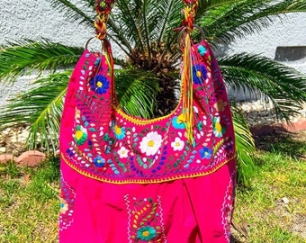 May include: A pink fabric tote bag with colorful embroidered flowers. The bag has a long, colorful braided strap.