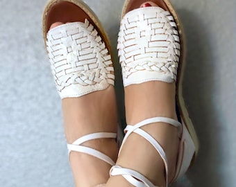 May include: White leather sandals with woven straps and laces that tie around the ankles.
