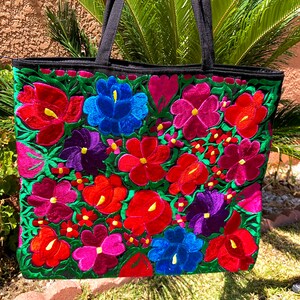 May include: A black tote bag with colorful embroidered flowers in shades of red, pink, blue, purple, and green. The flowers are arranged in a floral pattern on the front of the bag.