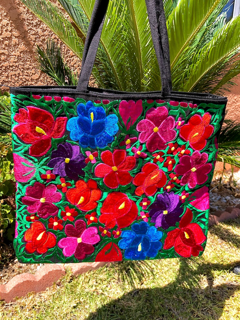 May include: A black tote bag with colorful embroidered flowers in shades of red, pink, blue, purple, and green. The flowers are arranged in a floral pattern on the front of the bag.