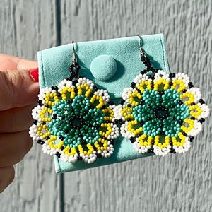 K&ouml;nnte beinhalten: Ein Paar Perlenohrringe mit Blumenmuster. Die Ohrringe sind aus schwarzen, wei&szlig;en, gelben und t&uuml;rkisfarbenen Perlen gefertigt. Die Ohrringe werden auf einem hellblauen Samtbeutel pr&auml;sentiert.