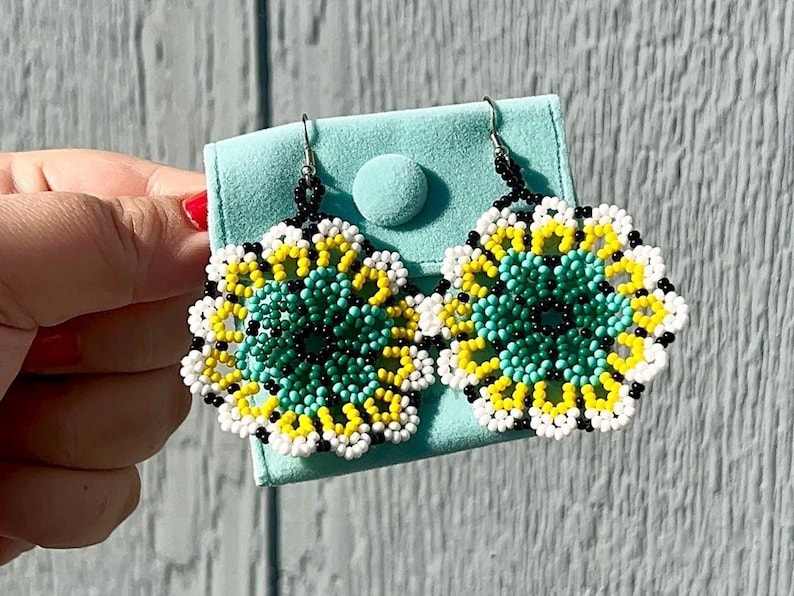 K&ouml;nnte beinhalten: Ein Paar Perlenohrringe mit Blumenmuster. Die Ohrringe sind aus schwarzen, wei&szlig;en, gelben und t&uuml;rkisfarbenen Perlen gefertigt. Die Ohrringe werden auf einem hellblauen Samtbeutel pr&auml;sentiert.