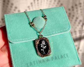 Peut inclure: Un collier en chaîne dorée avec un pendentif noir et or représentant une rose blanche. Le collier est dans une pochette en velours turquoise avec le texte "LATINA PALACE" imprimé dessus.