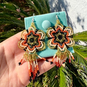 May include: A pair of beaded earrings with a floral design in shades of orange, black, gold, and white. The earrings have a tassel fringe.