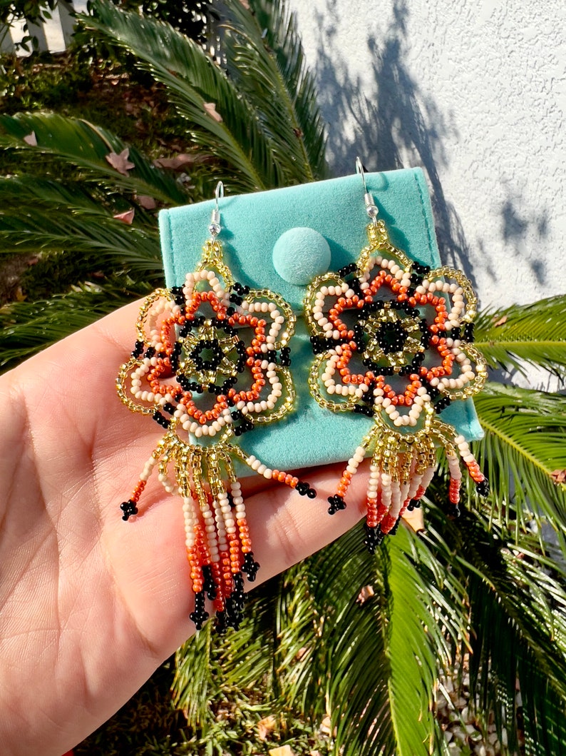May include: A pair of beaded earrings with a floral design in shades of orange, black, gold, and white. The earrings have a tassel fringe.