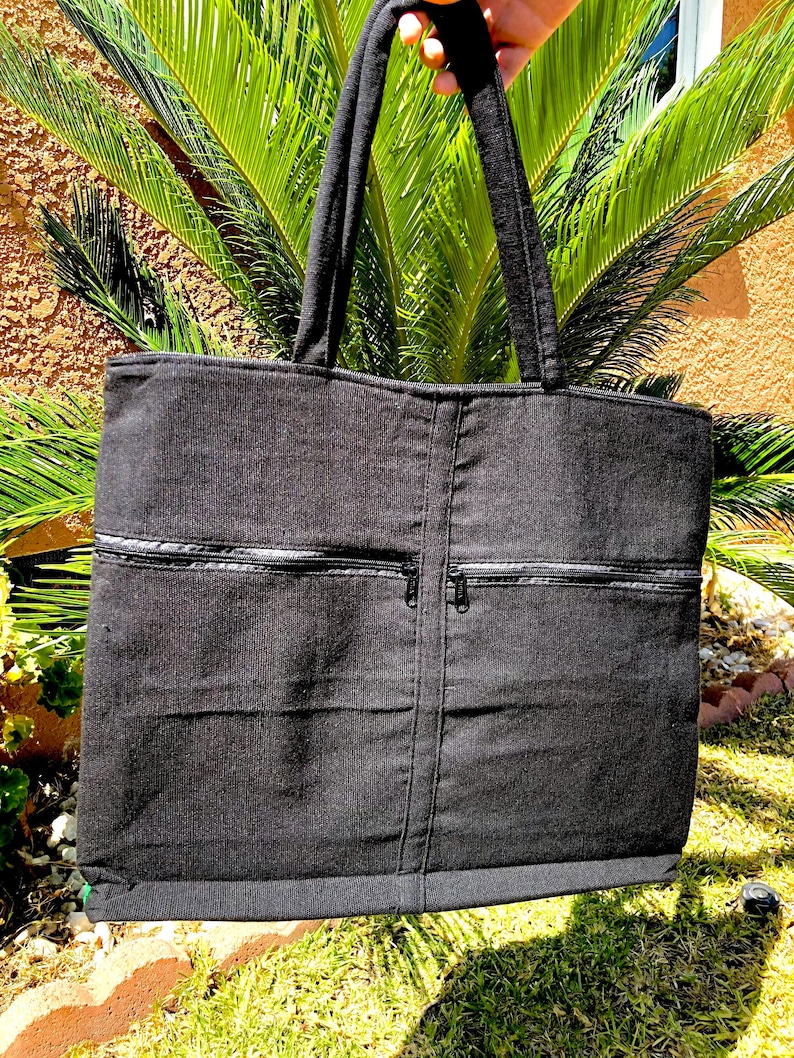 May include: Black canvas tote bag with two zippered pockets on the front. The bag has a black fabric handle.