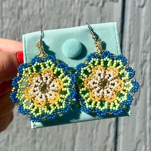 K&ouml;nnte beinhalten: Ein Paar Perlenohrringe mit einem Blumenmuster. Die Ohrringe sind aus blauen, gr&uuml;nen, gelben und wei&szlig;en Perlen gefertigt. Die Ohrringe h&auml;ngen an einem hellblauen Samtbeutel.