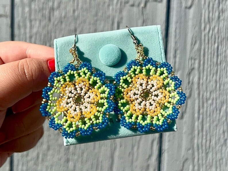 K&ouml;nnte beinhalten: Ein Paar Perlenohrringe mit einem Blumenmuster. Die Ohrringe sind aus blauen, gr&uuml;nen, gelben und wei&szlig;en Perlen gefertigt. Die Ohrringe h&auml;ngen an einem hellblauen Samtbeutel.