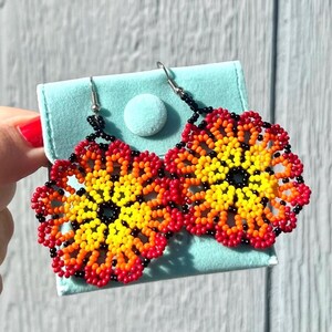 K&ouml;nnte beinhalten: Ein Paar Perlenohrringe in Form von Blumen. Die Ohrringe sind rot, orange, gelb und schwarz. Die Ohrringe h&auml;ngen an silbernen Haken.