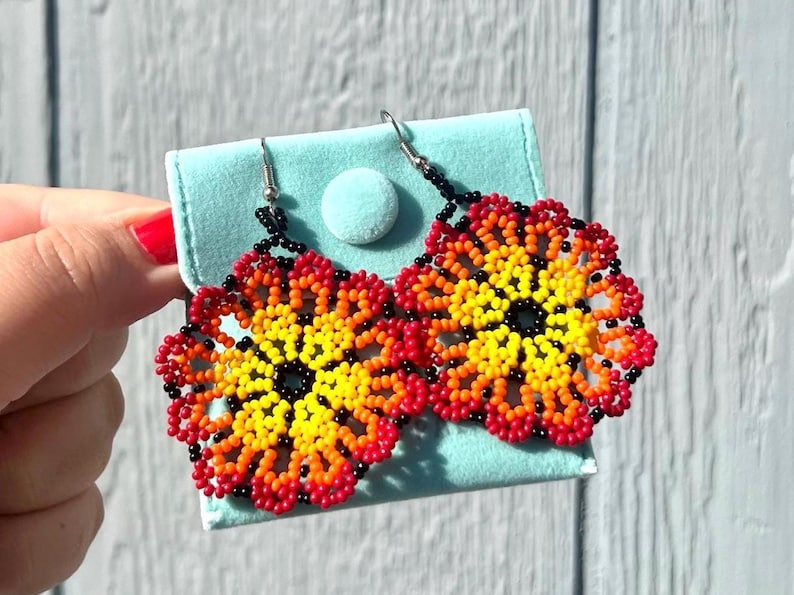 K&ouml;nnte beinhalten: Ein Paar Perlenohrringe in Form von Blumen. Die Ohrringe sind rot, orange, gelb und schwarz. Die Ohrringe h&auml;ngen an silbernen Haken.