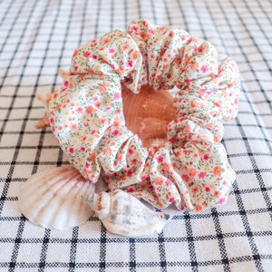 May include: A white fabric scrunchie with a floral pattern in shades of orange, pink, and yellow. The scrunchie is lying on a white and black checkered surface with two seashells.
