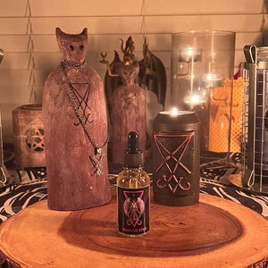 May include: A wooden figurine with a pentagram on its chest, a small bottle of oil labeled "Morning Star", and a black candle with a pentagram symbol on it. The items are arranged on a wooden slice, which is on a black and white patterned surface.