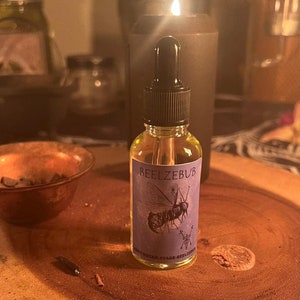 May include: A small glass bottle with a dropper, filled with a clear liquid. The bottle has a purple label with the text "BEELZEBUB" and a black and white illustration of a fly. The bottle is sitting on a wooden surface.