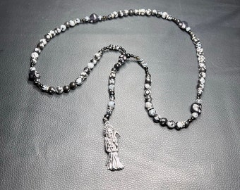 May include: A beaded necklace featuring black and white speckled beads and a silver pendant. The pendant depicts a robed figure with a scythe. The necklace is displayed on a black surface.