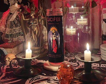 May include: A black candle holder with a lit candle sits on a black and white patterned table cloth. A glass jar candle with the label "LUCIFER MORNING STAR" is in the background. A red rose and a glass jar with tea lights are also visible.
