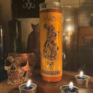 K&ouml;nnte beinhalten: Eine gelbe Kerze mit einer Schwarzwei&szlig;illustration eines D&auml;mons, der eine Trompete h&auml;lt. Die Kerze ist mit "MARBAS" beschriftet und hat den Text "VENERATISA-LIBERACA-BIASA-ICAR MARBAS" auf der Unterseite. Die Kerze ist von drei kleinen, brennenden Teelichtern auf einer Holzoberfl&auml;che umgeben.