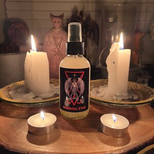 May include: A bottle of "Morning Star" spray with a black label featuring a winged figure, set on a wooden surface. Two large white candles and two tea lights are lit, creating a mystical ambiance. A pink statue is in the background.