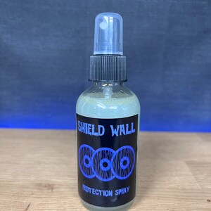 May include: A clear glass bottle with a black spray top and a black label that says "SHIELD WALL PROTECTION SPRAY". The label has a graphic of three blue interlocking circles with white dots.