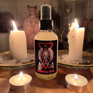 May include: A clear glass spray bottle with a black cap and a label featuring a winged figure and the words "MORNING STAR". The bottle is surrounded by lit candles and a statue.