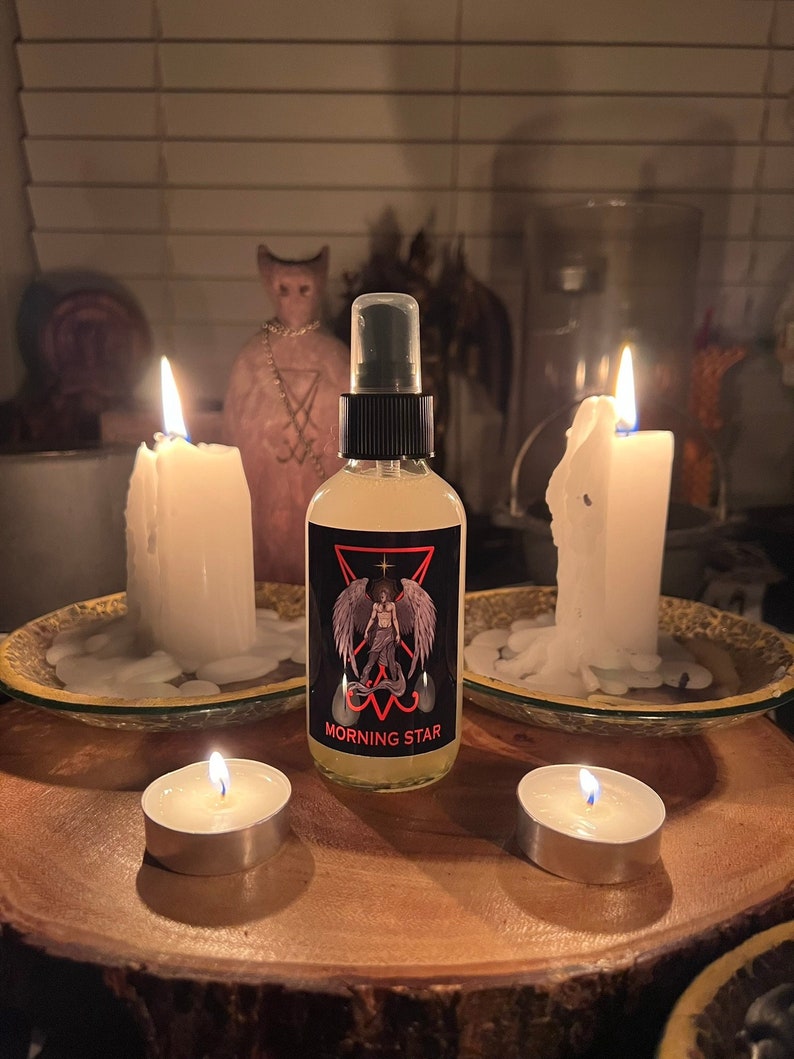 May include: A bottle of "Morning Star" spray with a black label featuring a winged figure, set on a wooden surface. Two large white candles and two tea lights are lit, creating a mystical ambiance. A pink statue is in the background.