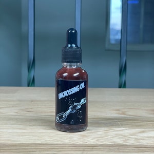 May include: A small brown glass bottle with a black dropper top. The bottle has a black label with white text that reads "UNCROSSING OIL". The label also features a graphic of a broken chain.
