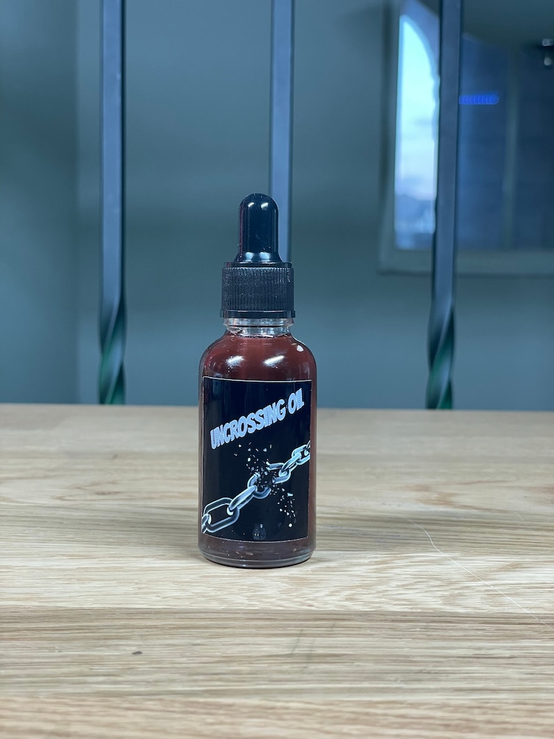 May include: A small brown glass bottle with a black dropper top. The bottle has a black label with white text that reads "UNCROSSING OIL". The label also features a graphic of a broken chain.