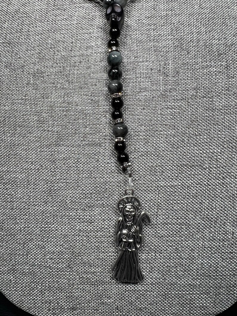 May include: A silver charm necklace with a black and grey beaded chain. The charm is a depiction of the Grim Reaper holding a scythe and a skull.