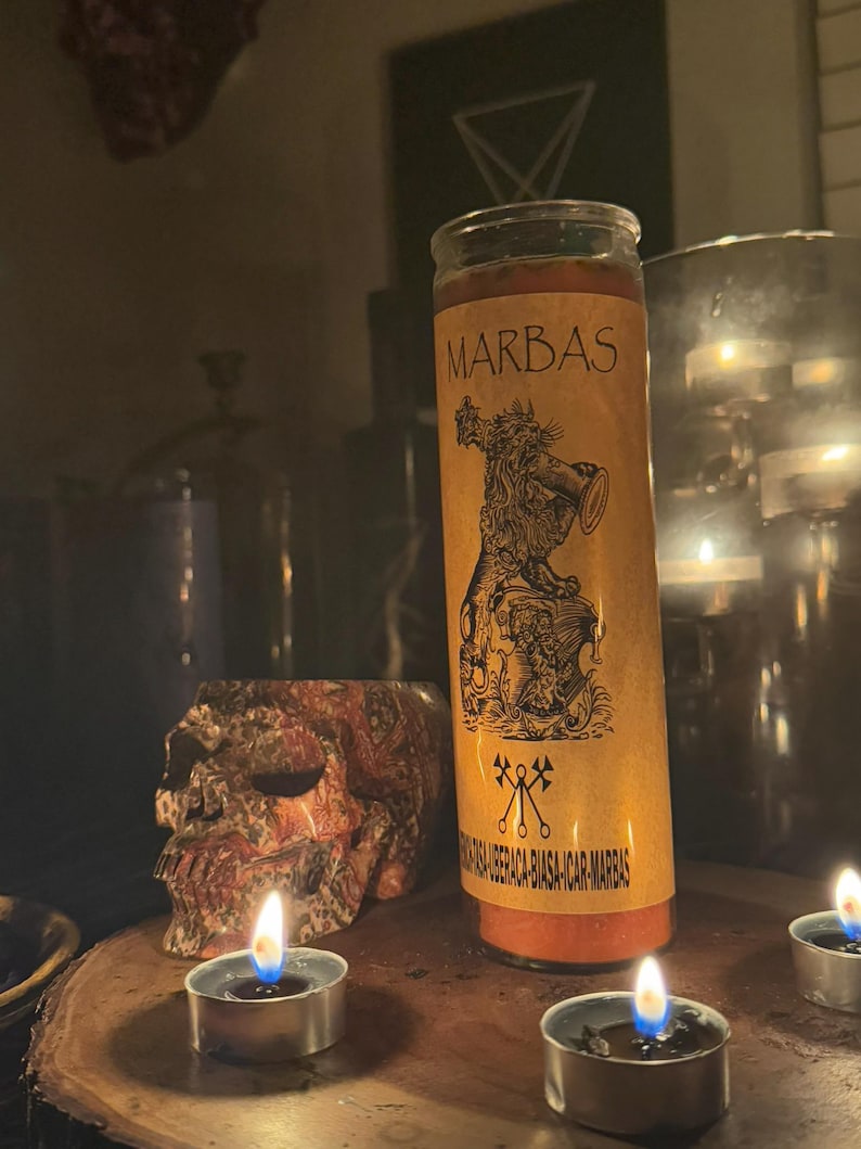 K&ouml;nnte beinhalten: Eine gelbe Kerze mit einem schwarzen Etikett, auf dem "MARBAS" steht, und einer Schwarzwei&szlig;illustration eines D&auml;mons, der eine Trompete h&auml;lt. Die Kerze ist von drei brennenden Teelichtern auf einer Holzoberfl&auml;che umgeben.