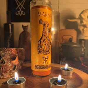 K&ouml;nnte beinhalten: Eine gelbe Kerze mit einem schwarz-wei&szlig;en Bild eines D&auml;mons mit dem Text "MARBAS" und "RENICH-TASA-UBERCACA-BIASA-HACA" auf dem Etikett. Die Kerze ist von drei kleinen, brennenden Teelichtern umgeben.