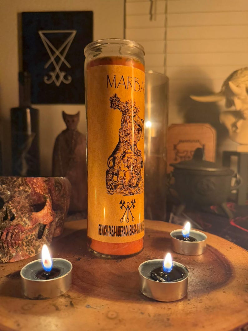 K&ouml;nnte beinhalten: Eine gelbe Kerze mit einem schwarz-wei&szlig;en Bild eines D&auml;mons mit dem Text "MARBAS" und "RENICH-TASA-UBERCACA-BIASA-HACA" auf dem Etikett. Die Kerze ist von drei kleinen, brennenden Teelichtern umgeben.
