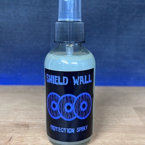 May include: A clear glass bottle with a black spray nozzle and a black label. The label says "Shield Wall" and features three blue and white shields with a white background. The label also says "Protection Spray".