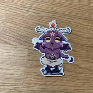 Pu&ograve; includere: Un piccolo diavolo di cartone animato viola con corna, ali e un pentagramma sulla testa. Il diavolo &egrave; in piedi su una roccia con una piccola fiamma sulla testa. Il diavolo indossa un pannolino bianco e ha un'espressione maliziosa sul viso.