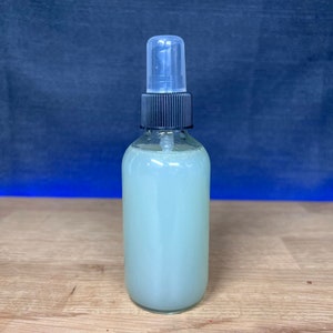 May include: A clear glass bottle with a black spray top filled with a light green liquid. The bottle is sitting on a light brown wooden surface.
