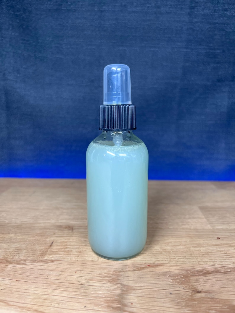 May include: A clear glass bottle with a black spray top filled with a light green liquid. The bottle is sitting on a light brown wooden surface.
