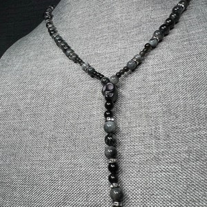 May include: A black and gray beaded necklace with a skull pendant. The beads are round and have a smooth, polished finish. The necklace is approximately 20 inches long.