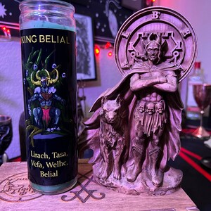 May include: A green candle with a black label that reads "King Belial" and "Lirach, Tasa, Vefa, Welhc, Belial". The candle is next to a wooden statue of a figure with a wolf at its feet. The statue is on a wooden box with a carved design.