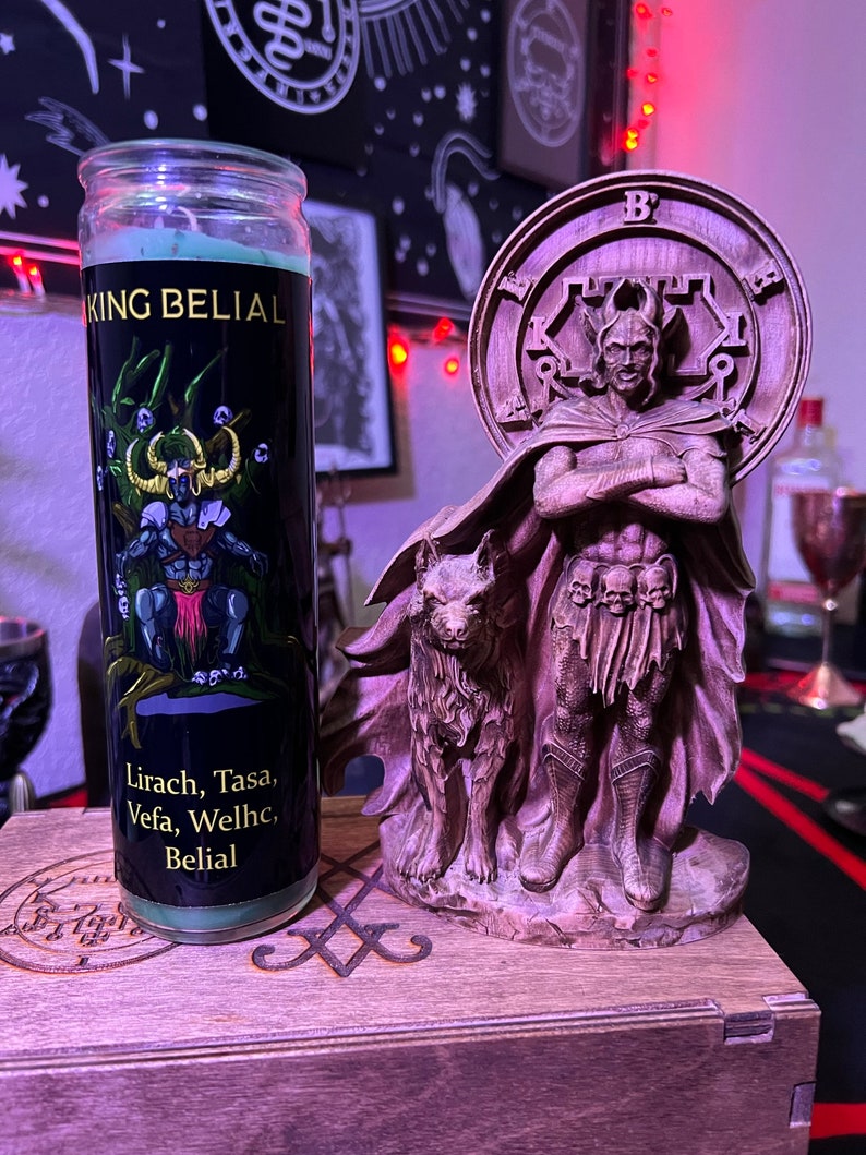 May include: A green candle with a black label that reads "King Belial" and "Lirach, Tasa, Vefa, Welhc, Belial". The candle is next to a wooden statue of a figure with a wolf at its feet. The statue is on a wooden box with a carved design.