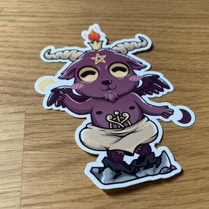 Pu&ograve; includere: Un piccolo diavolo di cartone animato viola e nero con corna, ali e un pentagramma sulla fronte. Il diavolo indossa un panno bianco e sta in piedi su una roccia. Lo sfondo &egrave; una superficie di legno.