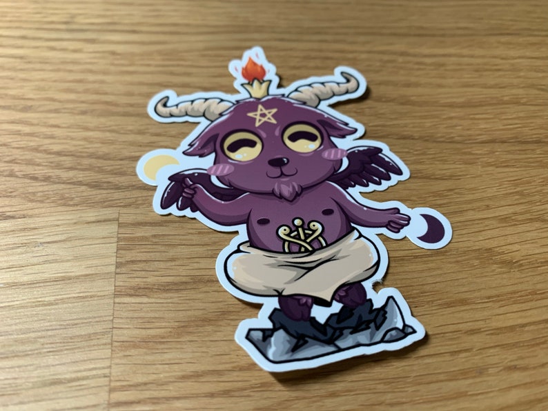 Pu&ograve; includere: Un piccolo diavolo di cartone animato viola e nero con corna, ali e un pentagramma sulla fronte. Il diavolo indossa un panno bianco e sta in piedi su una roccia. Lo sfondo &egrave; una superficie di legno.