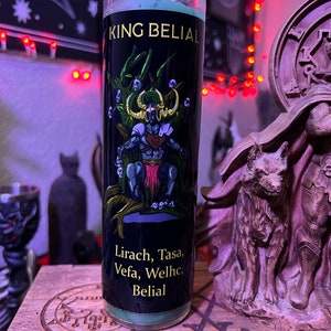 May include: A green glass candle with a black label. The label features a demon-like figure with a crown and a sword, surrounded by skulls. The text on the label reads "King Belial" and "Lirach, Tasa, Vefa, Welhc, Belial".