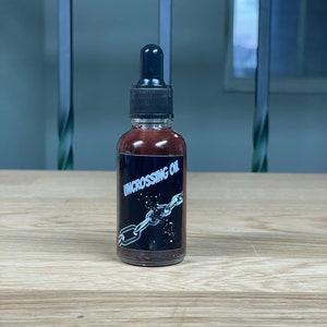 May include: A small brown glass bottle with a black dropper top. The bottle label is black with white text that reads "Uncrossing Oil" and a graphic of a broken chain.