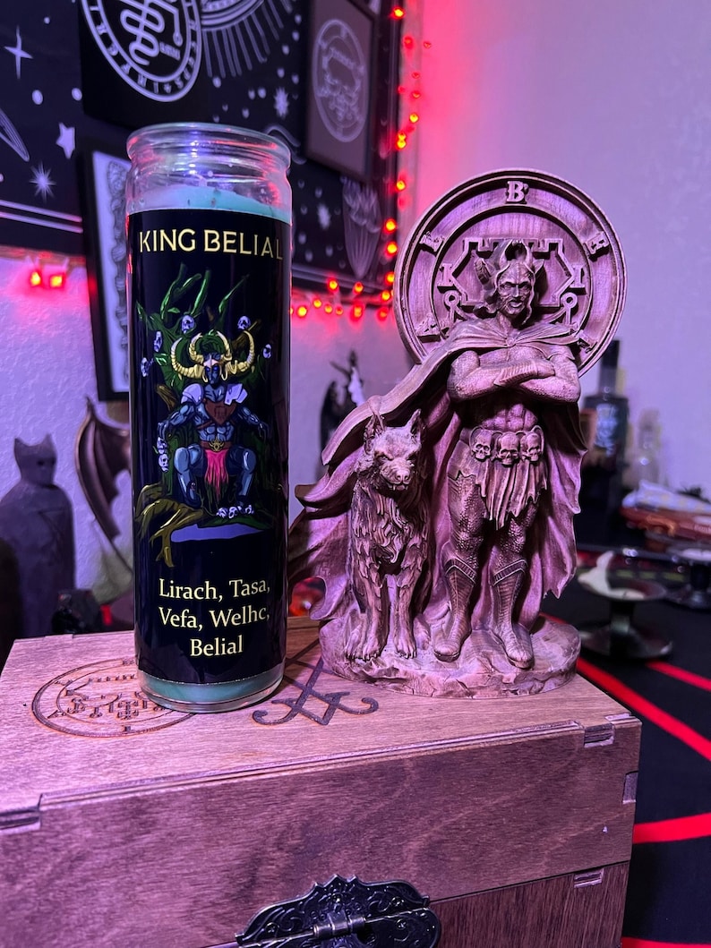 May include: A green candle with a black label that reads "King Belial" and "Lirach, Tasa, Vefa, Welhc, Belial". The candle is next to a wooden statue of a figure with a crown and a wolf at its feet.