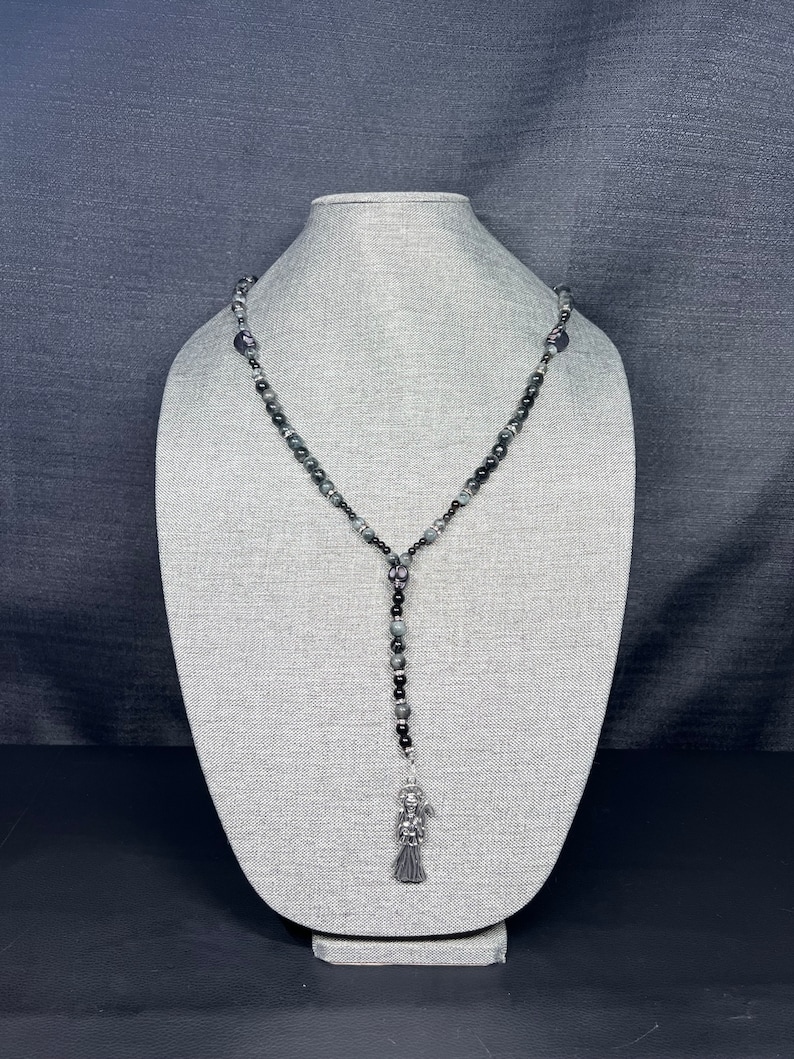 May include: A black and gray beaded necklace with a silver charm pendant. The charm is a figure of a woman with a long flowing dress and a crown.