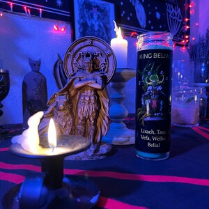 May include: A black and green candle with the text "KING BELIAL" and "Lirach, Tasa, Vefa, Welhe Belial" on the label. The candle is sitting on a table with a red and black striped tablecloth. There is a small white candle in a candlestick to the left of the candle.